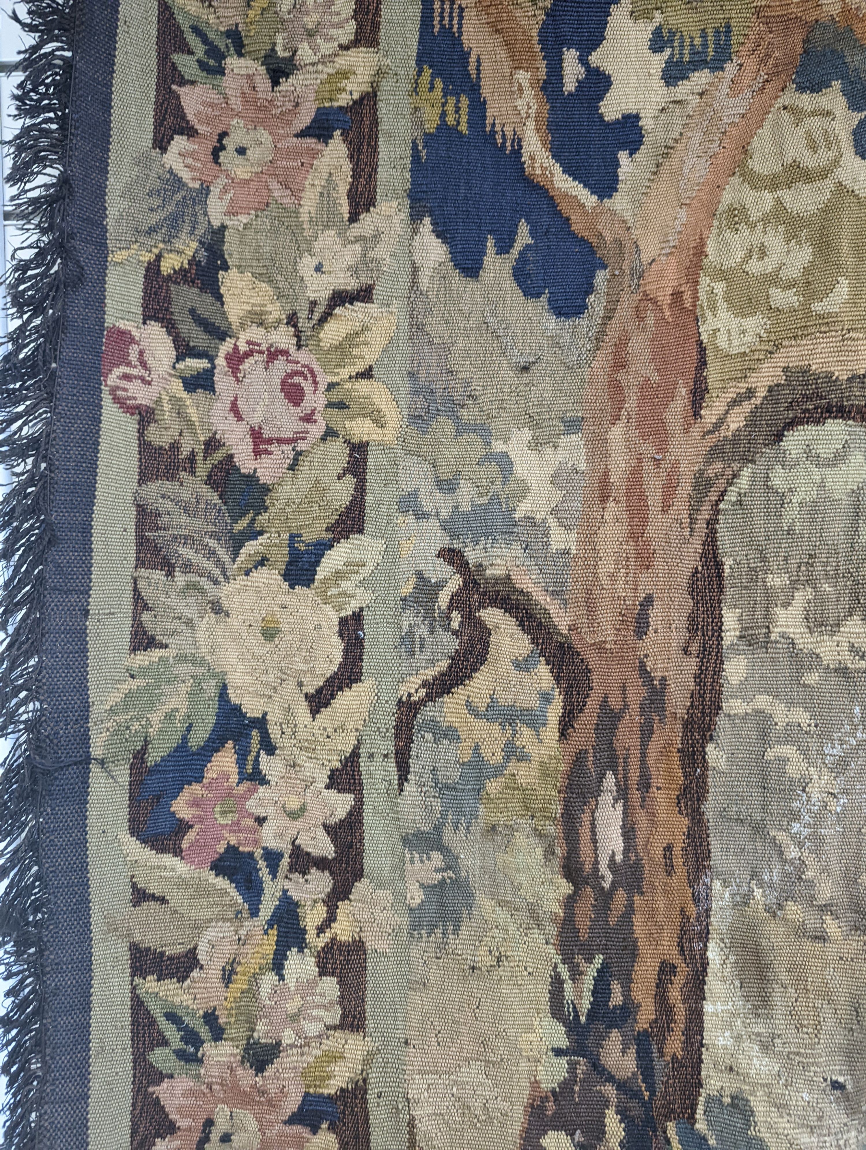 A late 19th / early 20th century French tapestry depicting a woodland scene with trees and flowers with a floral border woven in autumnal colours